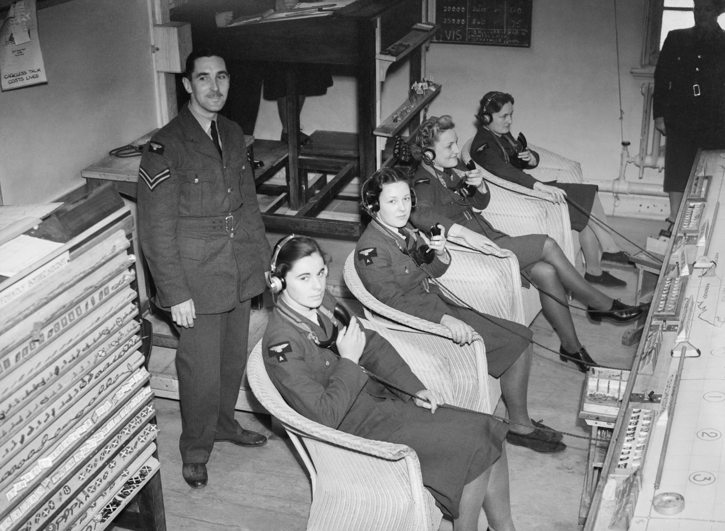 Women in uniform wearing telephone headsets sit at a long desk taking down reports in a wartime operations room