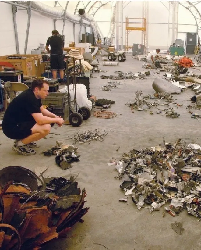 Elon Musk crouches on a hangar floor looking at piles of twisted, scorched rocket debris laid out in rows