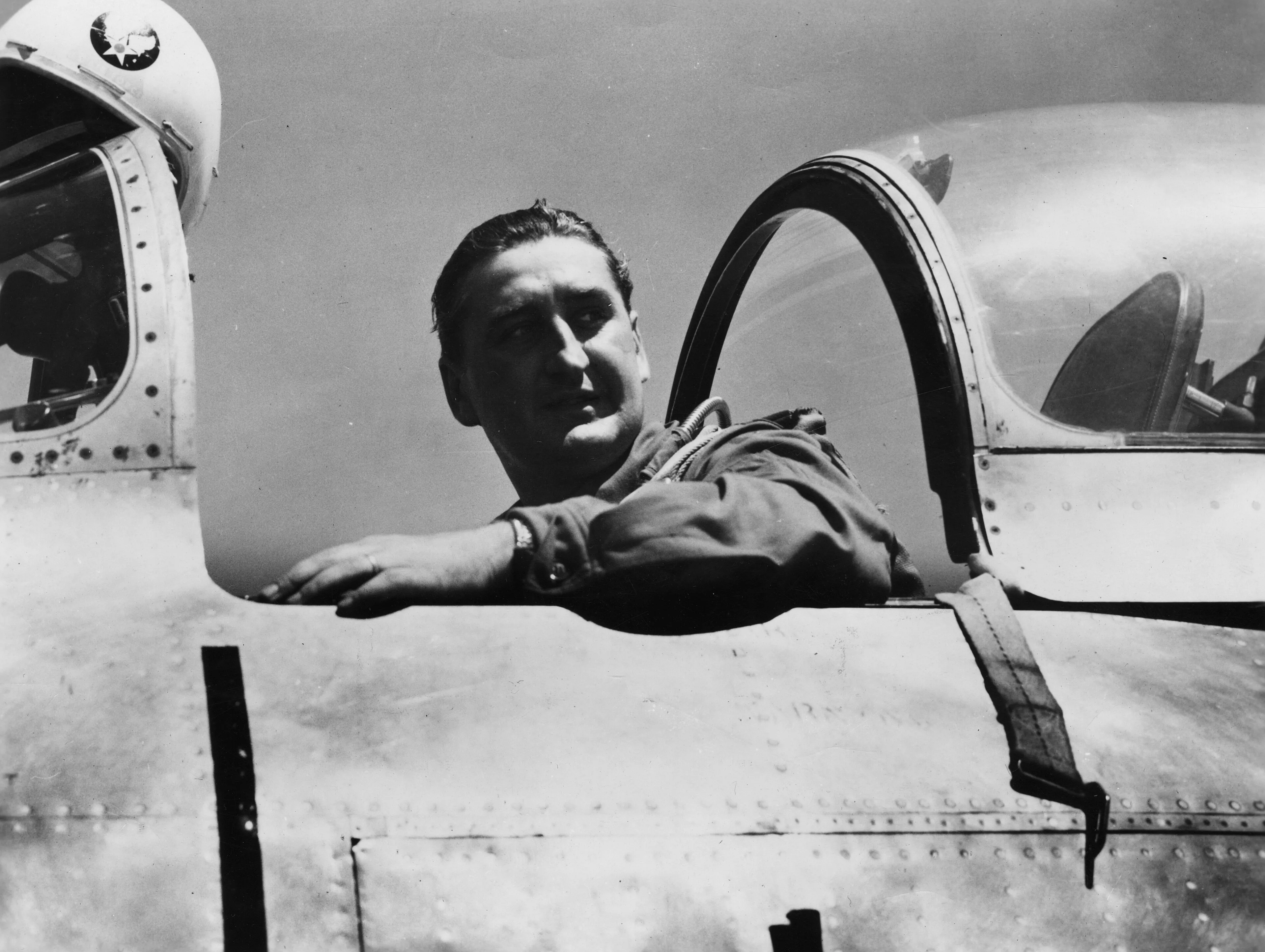 A pilot in a flying helmet sits in the open cockpit of a jet fighter