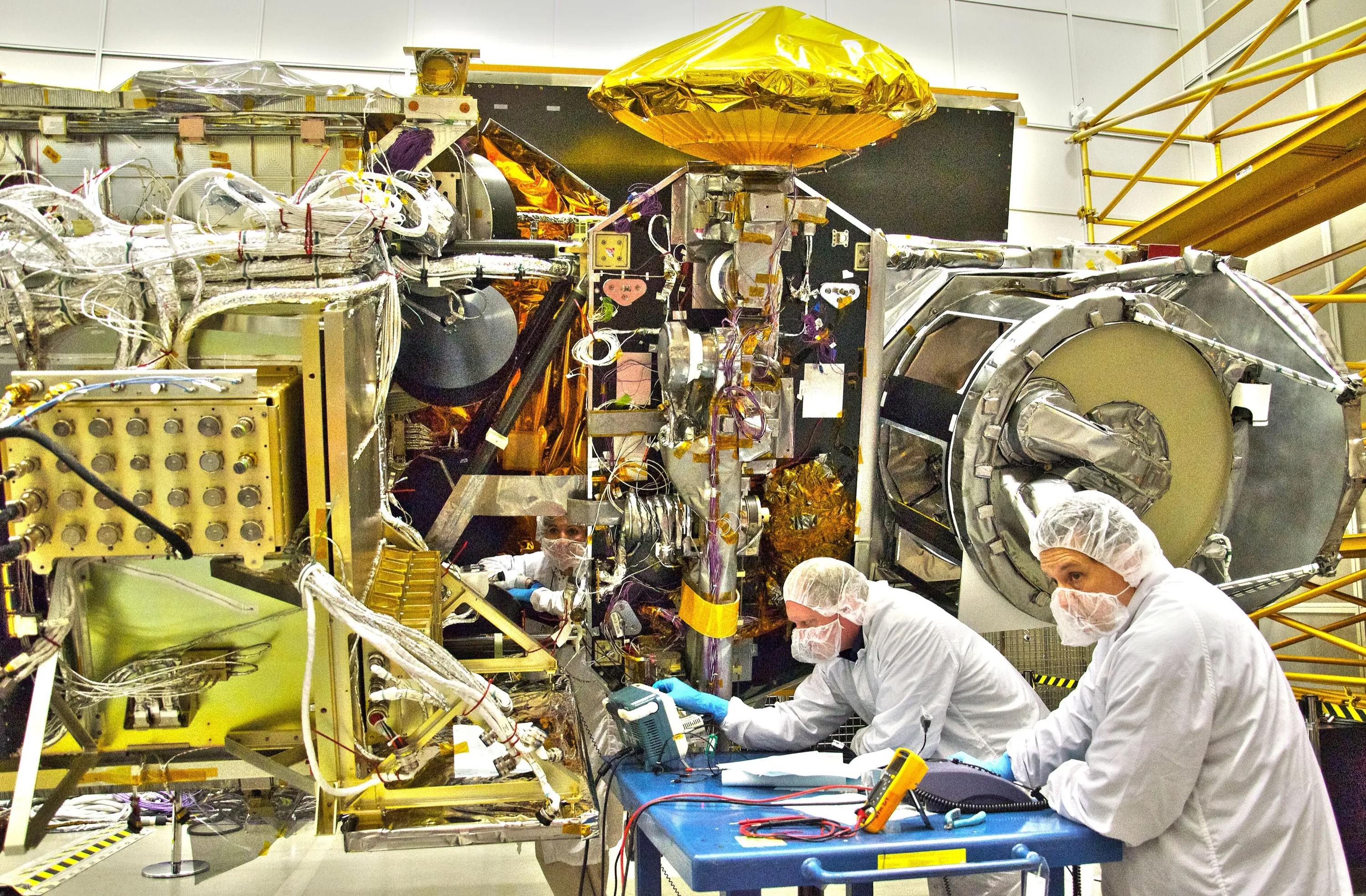 Three engineers in white clean-room suits and hoods working on a large satellite frame wrapped in gold foil