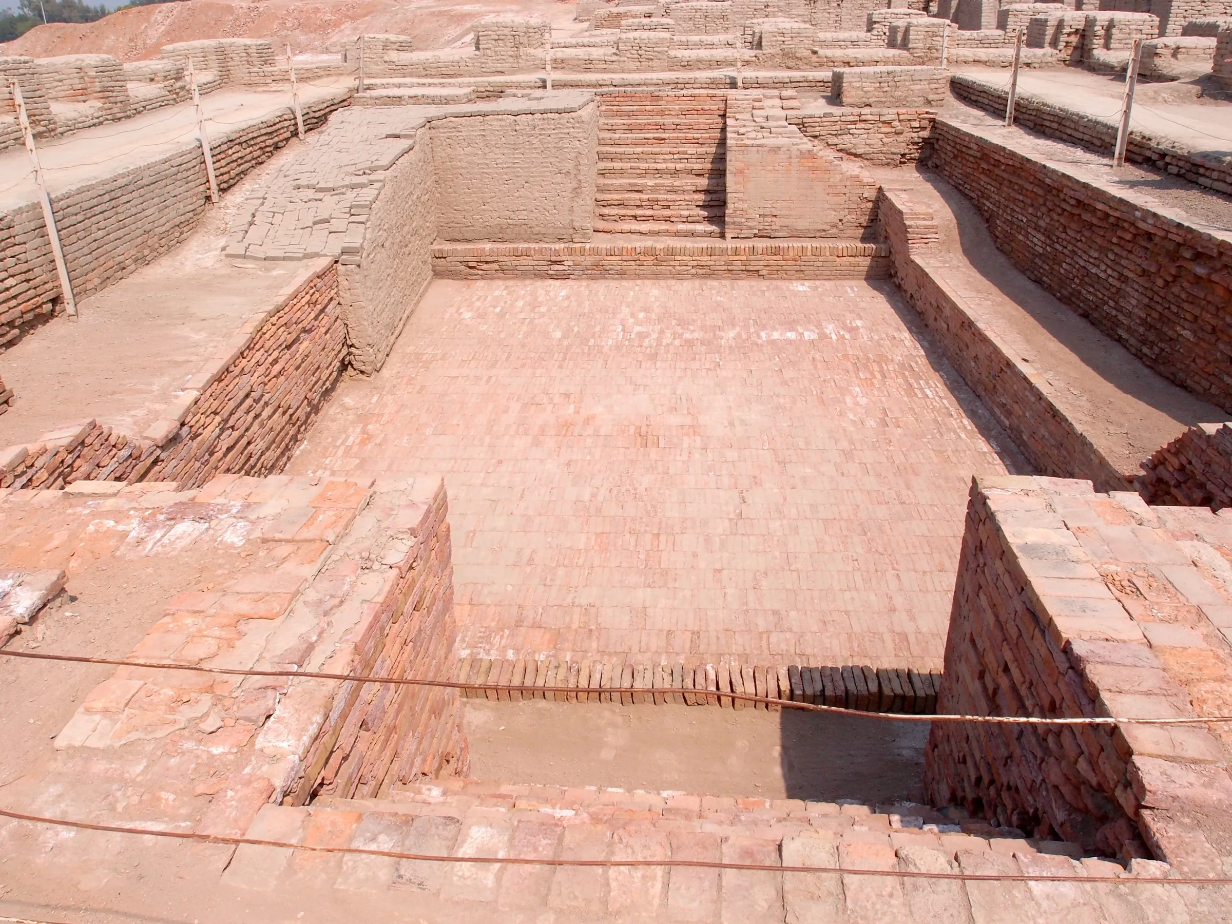 An excavated rectangular brick tank with steps leading down at each end, surrounded by brick walls and ruined rooms