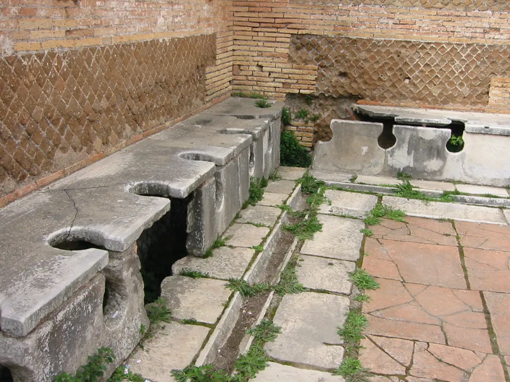A row of stone latrine seats with keyhole-shaped openings along a wall in the ruins of Ostia, the port of ancient Rome