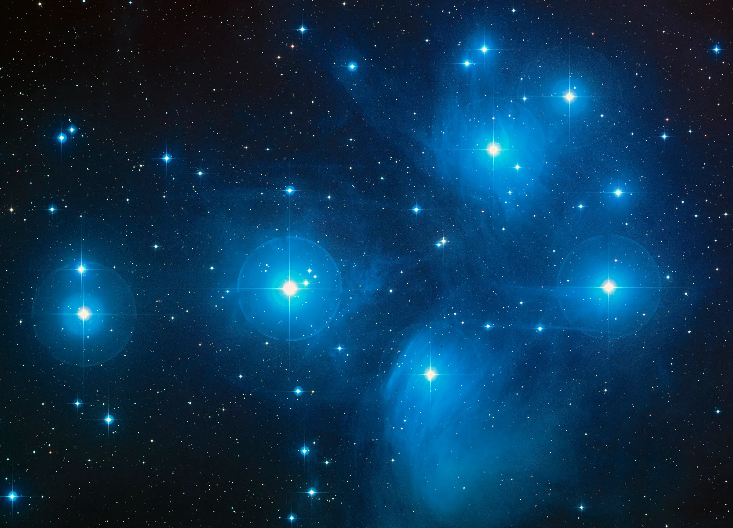 A telescope photograph of the Pleiades star cluster: a handful of bright blue-white stars wrapped in faint blue nebulosity against a black sky