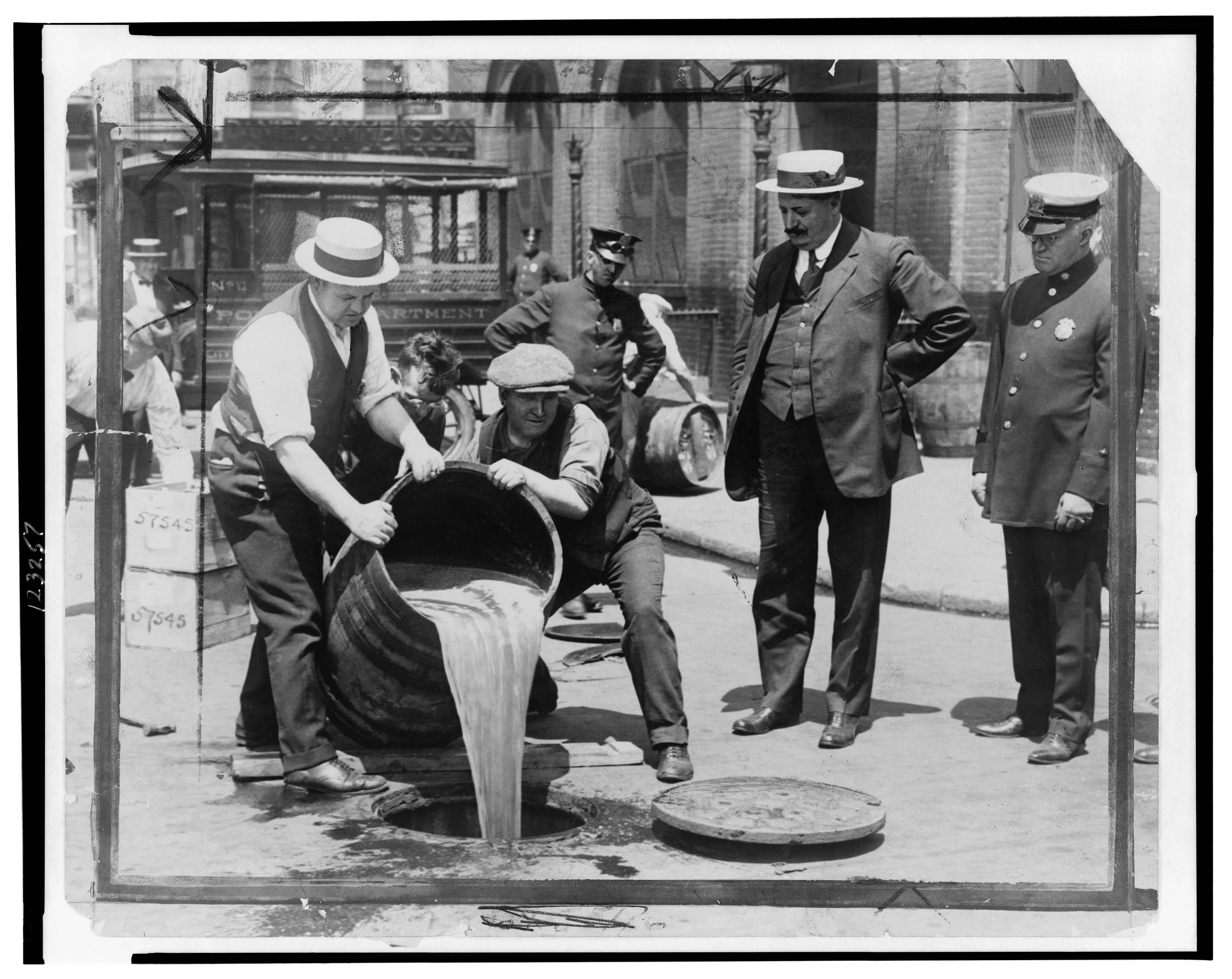 Black-and-white photograph of men in suits and hats watching agents pour liquor from barrels into a street sewer
