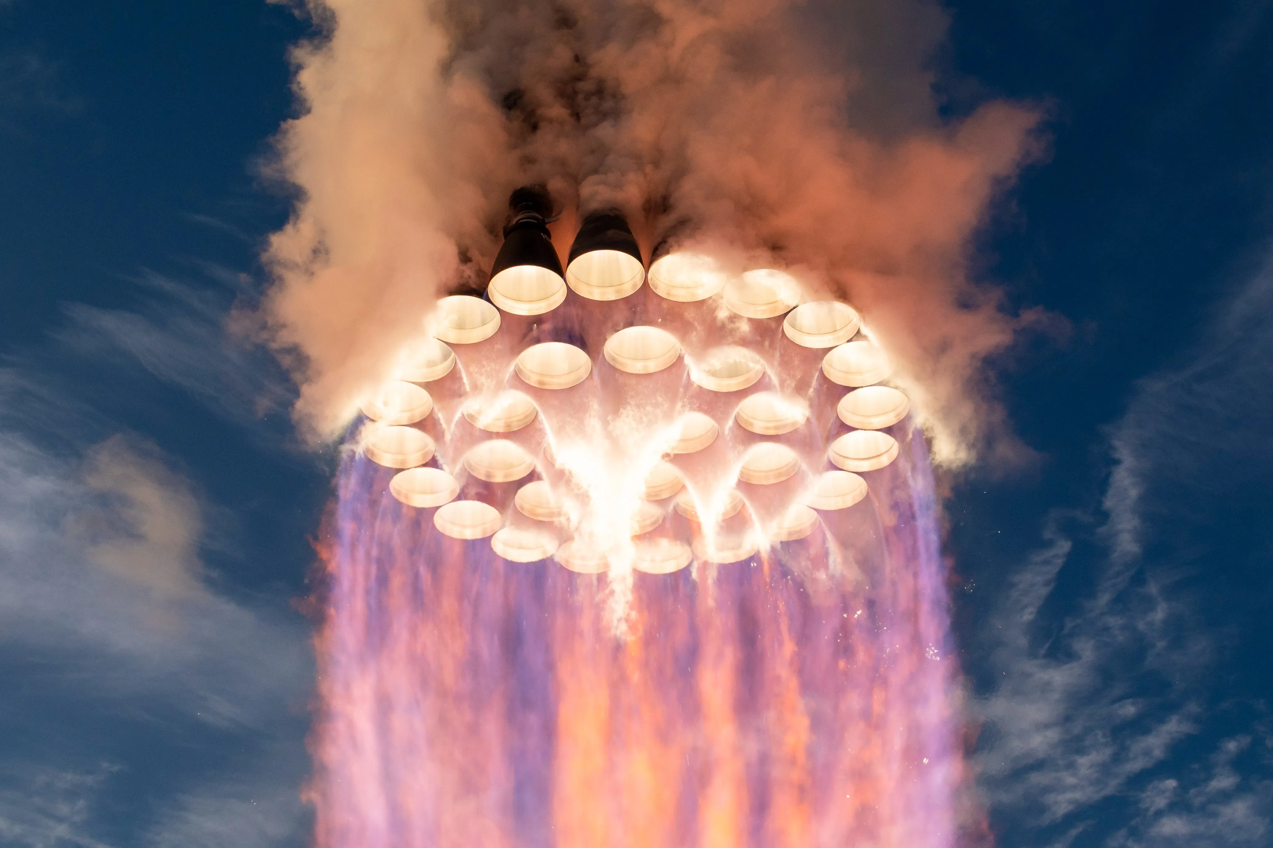 Looking up at the base of a booster in flight: a ring of glowing Raptor engine bells inside a wider ring, firing violet flame against a blue sky