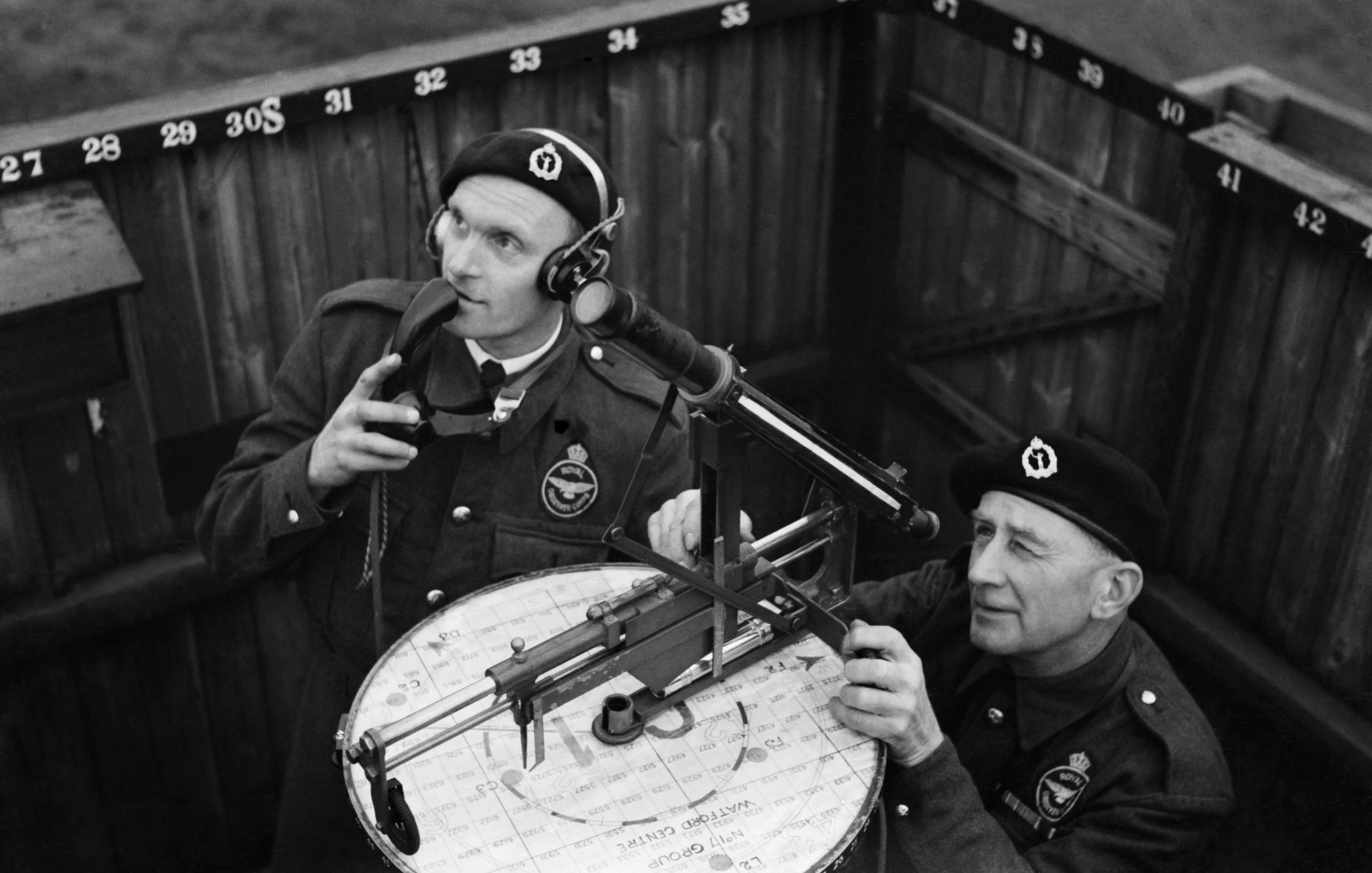 Two men at an outdoor observation post: one wears a telephone headset on his chest and speaks into it, the other works a sighting instrument on a plotting table