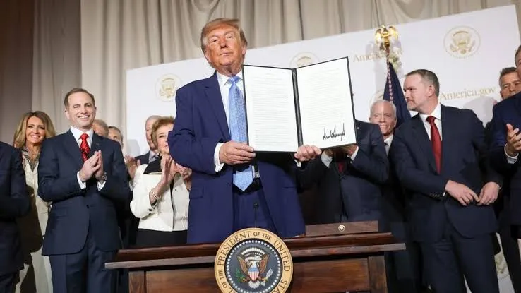 Donald Trump holds open a folder showing a signed document, standing at a lectern with the presidential seal; officials applaud behind him in front of a backdrop printed with America.gov