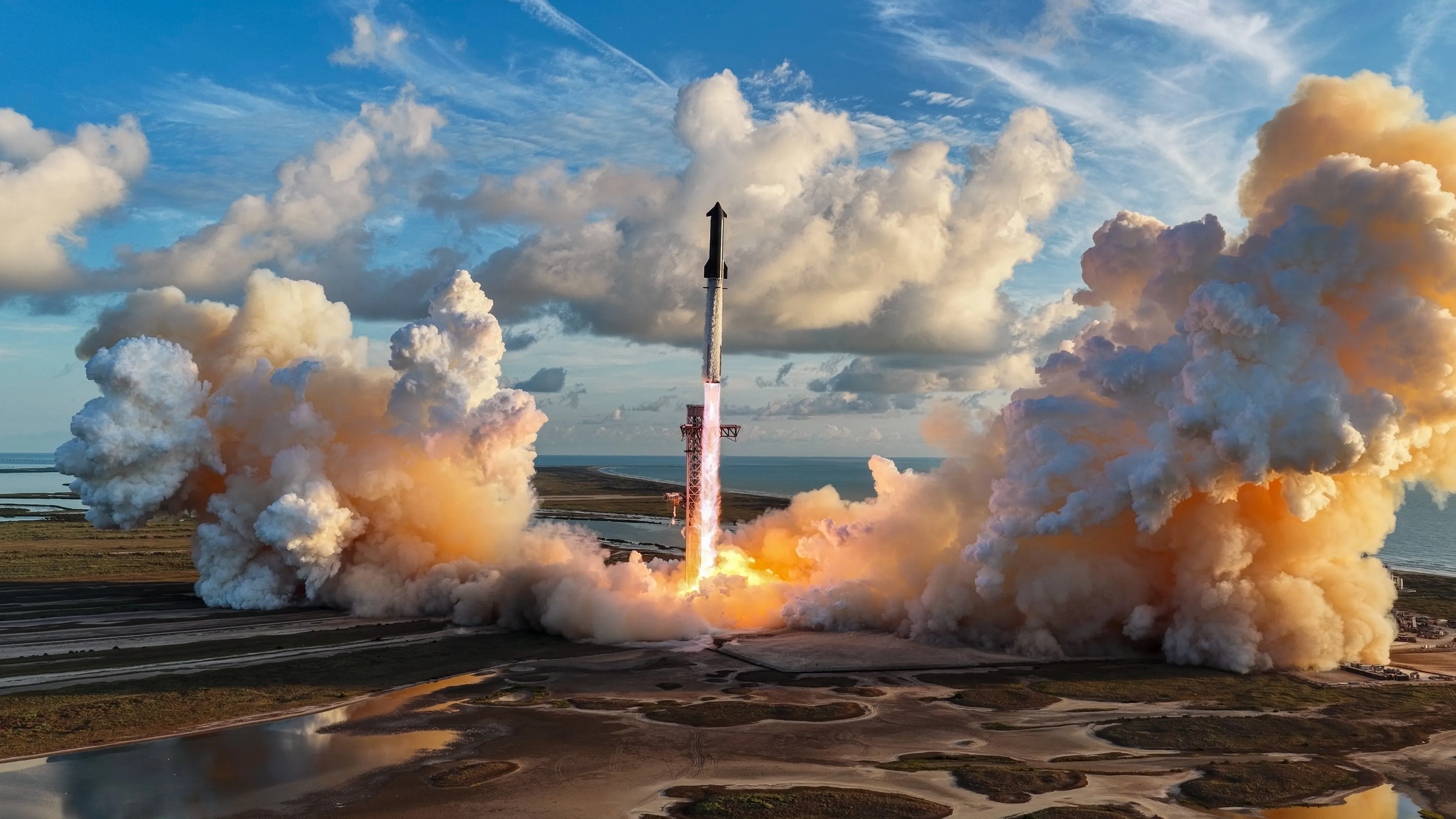 Starship climbs on a column of flame from the launch site, with huge billows of exhaust spreading across marshy ground and the sea behind