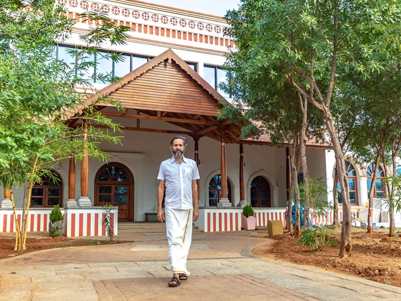 Sridhar Vembu, in a white shirt and white veshti with sandals, walking along a stone path away from a white building with a pitched wooden porch roof, arched windows and young trees in front
