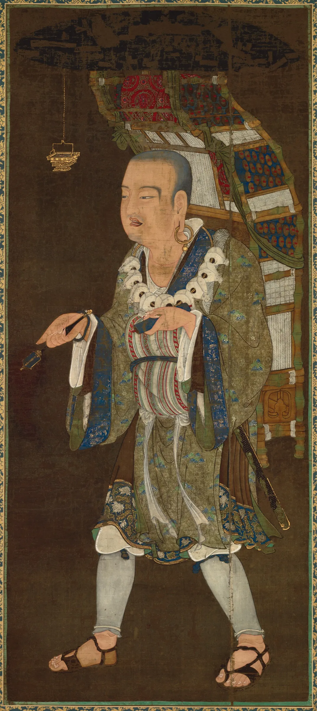 A Japanese hanging scroll painting of a Buddhist monk walking, carrying a tall frame pack loaded with scrolls on his back